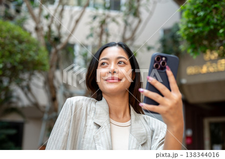 Pretty asian woman holding phone looking away while waiting for taxi or delivery in front of a condo Pretty asian woman holding phone looking away while waiting for taxi or delivery in front of a condo 138344016