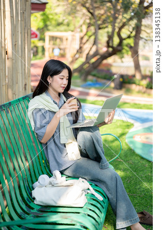 Pretty asian woman holding coffee watching on laptop while sitting knee up on bench in garden park. 138345138