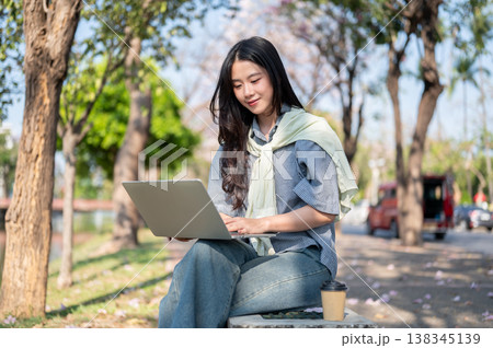 Pretty asian woman looking at a laptop over crossed legs while sitting on concrete bench at sidewalk 138345139