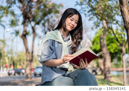 Pretty asian woman reading a book holding over crossed legs as sitting on bench at sidewalk by road. Pretty asian woman reading a book holding over crossed legs as sitting on bench at sidewalk by road. 138345141