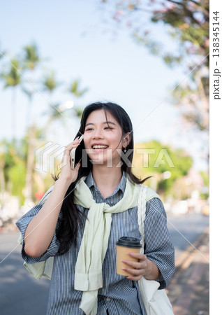 Smiling asian woman holding coffee cup talking on phone call while standing or walking by the road. Smiling asian woman holding coffee cup talking on phone call while standing or walking by the road. 138345144