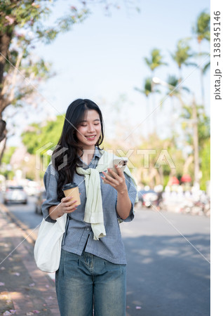 Happy asian woman holding coffee cup and looking at phone while walking on sidewalk by the city road Happy asian woman holding coffee cup and looking at phone while walking on sidewalk by the city road 138345146