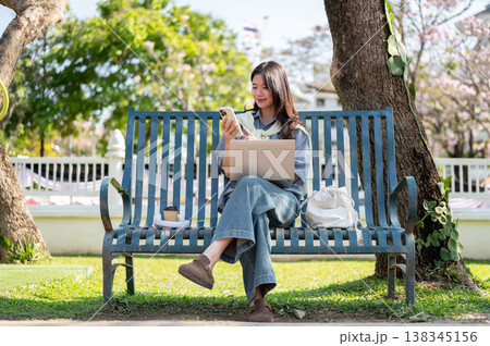 Pretty asian woman looking at phone over laptop while sitting cross legged on bench in garden park. Pretty asian woman looking at phone over laptop while sitting cross legged on bench in garden park. 138345156