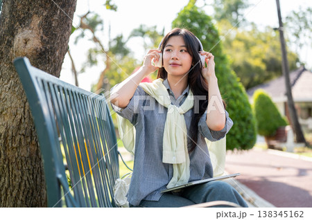 Pretty asian woman holding headphones listening to music while sitting on metal bench in garden park Pretty asian woman holding headphones listening to music while sitting on metal bench in garden park 138345162