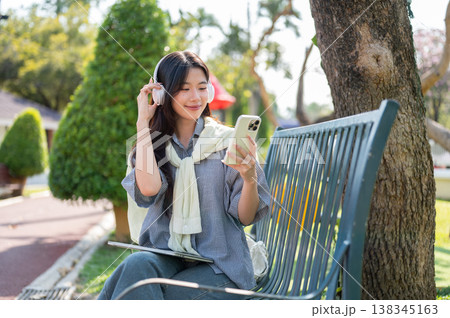 Pretty asian woman listening to music looking at phone while sitting on metal bench in garden park. 138345163