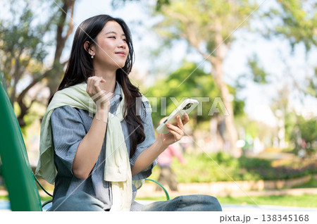 Pretty asian woman holding phone looking ahead while sitting folding legs on bench in garden or park 138345168