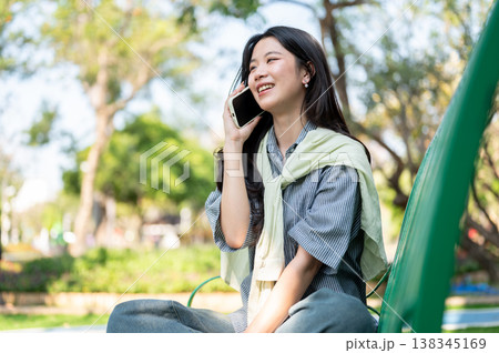 Pretty asian woman smiling as talking on phone while sitting folding legs on bench in garden or park 138345169