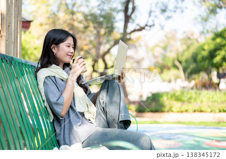 Pretty asian woman drinking coffee watching on laptop while sitting knee up on bench in garden park. Pretty asian woman drinking coffee watching on laptop while sitting knee up on bench in garden park. 138345172