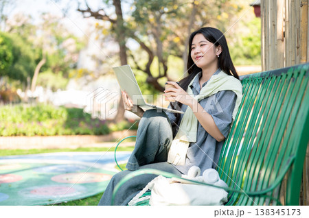 Pretty asian woman holding coffee cup looking away from laptop while sitting on bench in garden park Pretty asian woman holding coffee cup looking away from laptop while sitting on bench in garden park 138345173