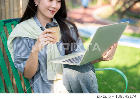Pretty asian woman holding coffee cup and looking at a laptop while sitting on bench in garden park. Pretty asian woman holding coffee cup and looking at a laptop while sitting on bench in garden park. 138345174