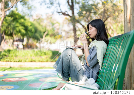 Pretty asian woman drinking coffee looking at laptop on knees while sitting on bench in garden park. Pretty asian woman drinking coffee looking at laptop on knees while sitting on bench in garden park. 138345175