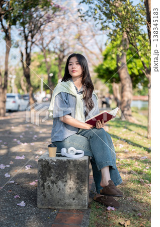Pretty asian woman looking away from a book while sitting cross legged on concrete bench at sidewalk Pretty asian woman looking away from a book while sitting cross legged on concrete bench at sidewalk 138345183