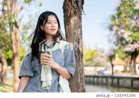 Pretty asian woman holding coffee enjoy the scenery while standing leaning on a tree by city's canal 138345200