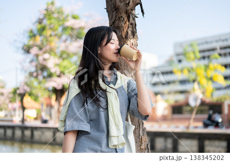 Pretty asian woman closing eyes enjoy drinking coffee while standing leaning on tree by city's canal Pretty asian woman closing eyes enjoy drinking coffee while standing leaning on tree by city's canal 138345202