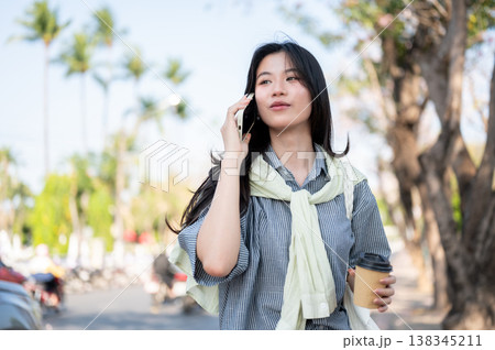 Pretty asian woman holding coffee taking or making a phone call while standing or walking by a road. 138345211