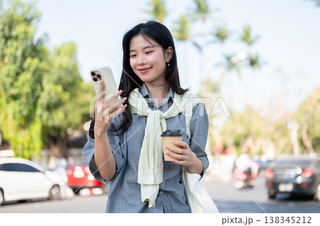 Smiling asian woman holding coffee cup looking at smartphone while standing or walking by the road. 138345212
