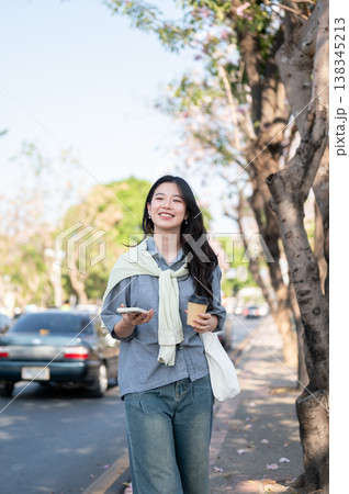 Asian woman smiling happily as holding coffee cup and phone while walking on sidewalk by the road. 138345213