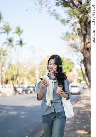 Pretty asian woman holding coffee cup as looking at phone while walking on sidewalk by the city road Pretty asian woman holding coffee cup as looking at phone while walking on sidewalk by the city road 138345214