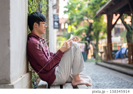 Glasses young asian man reading a book while sitting knee up leaning on concrete wall of a building. 138345914
