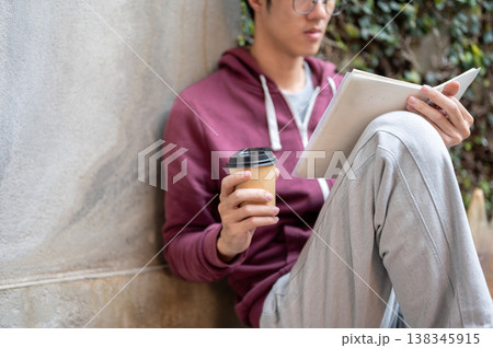 Glasses young asian man holding coffee reading a book while sitting knee up leaning on concrete wall Glasses young asian man holding coffee reading a book while sitting knee up leaning on concrete wall 138345915