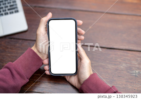 Top view of a man's hands holding white screen smartphone aside laptop while sitting at wooden table 138345923