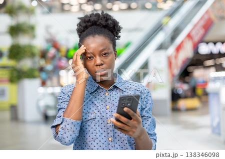 Young woman standing in a shopping mall, holding a mobile phone and touching her head, expressing feelings of headache, stress, and frustration from bad news or a difficult situation Young woman standing in a shopping mall, holding a mobile phone and touching her head, expressing feelings of headache, stress, and frustration from bad news or a difficult situation 138346098