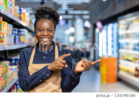 Friendly supermarket worker smiling, wearing an apron, and pointing fingers to the right, presenting a product or inviting customers to a specific area in a grocery store Friendly supermarket worker smiling, wearing an apron, and pointing fingers to the right, presenting a product or inviting customers to a specific area in a grocery store 138346166