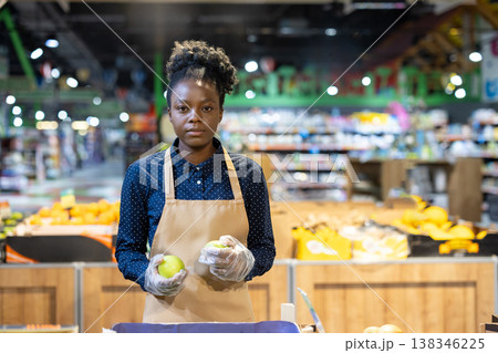 African american woman wearing an apron and gloves, holding green apples while working in the produce section of a grocery store, representing retail and customer service 138346225
