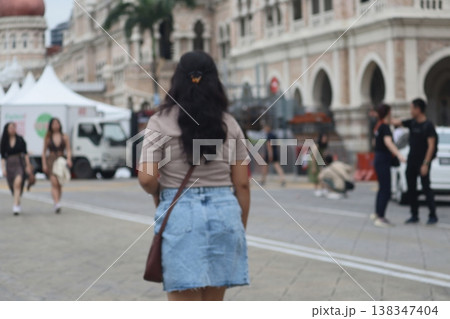 Woman walking through a public square in Kuala Lumpur, Malaysia, with colonial-era buildings and daily city life captured from a candid rear perspective. Woman walking through a public square in Kuala Lumpur, Malaysia, with colonial-era buildings and daily city life captured from a candid rear perspective. 138347404