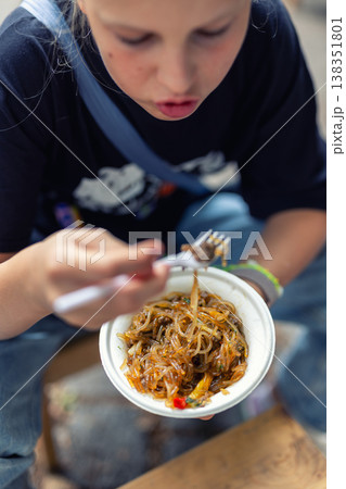 Candid portrait child eating noodles from bowl during streetfood event showing focus curiosity enjoyment casual lifestyle. Outdoor urban scene street food festival people taste international cuisine. Candid portrait child eating noodles from bowl during streetfood event showing focus curiosity enjoyment casual lifestyle. Outdoor urban scene street food festival people taste international cuisine. 138351801