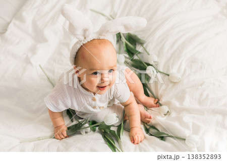 Cute funny baby with bunny ears, with a tulips at home on a bed of white bedding. Easter greeting card Cute funny baby with bunny ears, with a tulips at home on a bed of white bedding. Easter greeting card 138352893