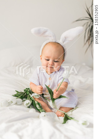 Cute baby with rabbit ears on his head and tulips at home on the bed. Easter and spring time concept. Cute baby with rabbit ears on his head and tulips at home on the bed. Easter and spring time concept. 138352905