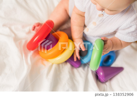 Close-up baby playing with colorful rainbow toy pyramid on white sunny bedroom. Toys for little kids. Child with educational toy. Close-up baby playing with colorful rainbow toy pyramid on white sunny bedroom. Toys for little kids. Child with educational toy. 138352906