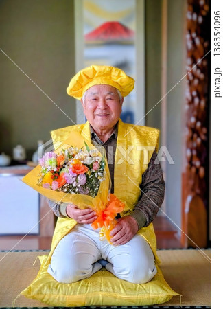 和室で傘寿を祝う黄色の祝い衣装を着た笑顔の高齢男性・花束を持つ記念写真 138354096