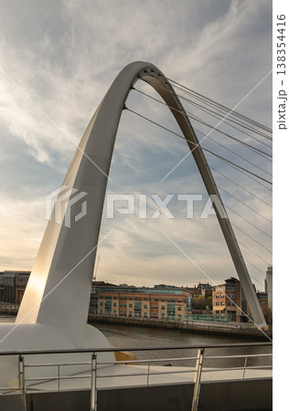 The sweeping arc of the Gateshead Millennium Bridge dominates this vertical frame, its pristine white steel slicing through a sky of soft, streaked clouds. Often called the "Blinking Eye," this 138354416
