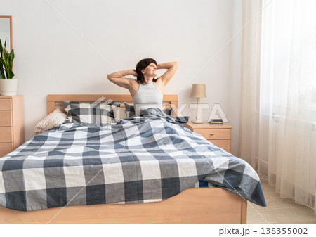 An adult woman feeling refreshed and well-rested, stretching her arms and waking up to natural light in a cozy bedroom. Authentic lifestyle 138355002