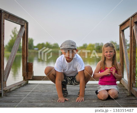A cute small boy with a flat cap and a sweet little girl are sitting on a wooden pier on the shore of a lake. Small kids are sitting on a pier at the edge of a pond. The concept of a carefree 138355487