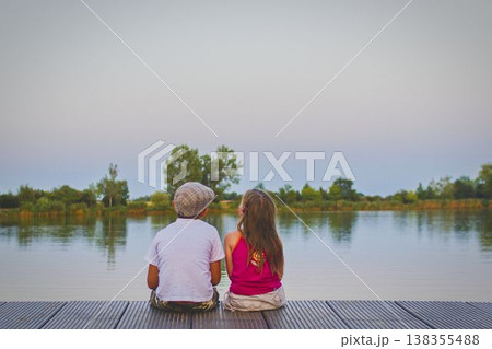 A little boy wearing a flat cap and a cute little girl sit with their backs turned on a wooden pier on the shore of a lake. Small children sit on a pier at the edge of a pond. The concept of a A little boy wearing a flat cap and a cute little girl sit with their backs turned on a wooden pier on the shore of a lake. Small children sit on a pier at the edge of a pond. The concept of a 138355488