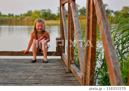 Young girl in swimsuit and shoes sitting on a wooden pier by the lake, smiling 138355570