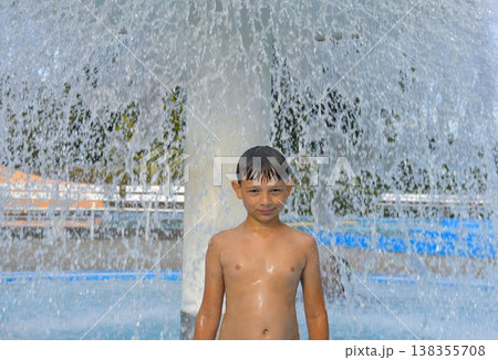 A boy under a waterfall at a public swimming pool. A little boy under a sprinkler at a swimming pool. Copy space. The concept of summer, summer holidays, free time, and relaxation. A boy under a waterfall at a public swimming pool. A little boy under a sprinkler at a swimming pool. Copy space. The concept of summer, summer holidays, free time, and relaxation. 138355708