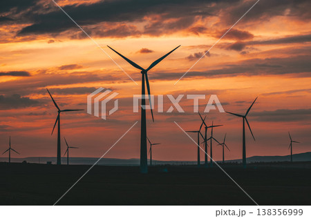Multiple wind turbines silhouetted against a vibrant sunset sky. The turbines stand tall against the backdrop of orange and red clouds 138356999