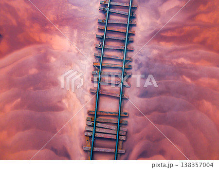 Aerial of a railroad tracks crossing a pink-hued lake. The perspective offers a unique view of the railway lines submerged in the shallow waters of the lake. Burlinskoe Lake, located in Bursol. 138357004