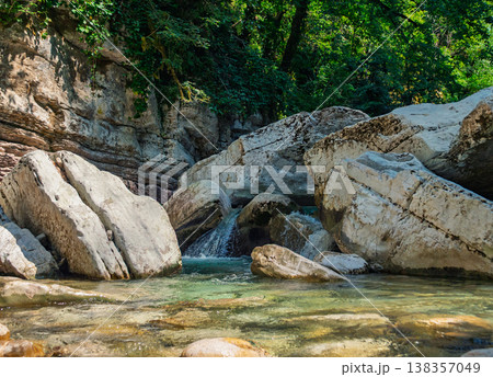 Flowing mountain river with clear water winding through a sunlit rocky gorge, surrounded by lush green trees and ancient stone formations Flowing mountain river with clear water winding through a sunlit rocky gorge, surrounded by lush green trees and ancient stone formations 138357049