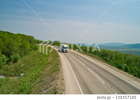 Aerial view truck travels along a winding highway in a rural area with green hills under clear skies Aerial view truck travels along a winding highway in a rural area with green hills under clear skies 138357185
