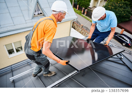 Workers building solar panel system on roof of house. Two men installers in helmets carrying photovoltaic solar module outdoors. Alternative, green and renewable energy generation concept. Workers building solar panel system on roof of house. Two men installers in helmets carrying photovoltaic solar module outdoors. Alternative, green and renewable energy generation concept. 138357696