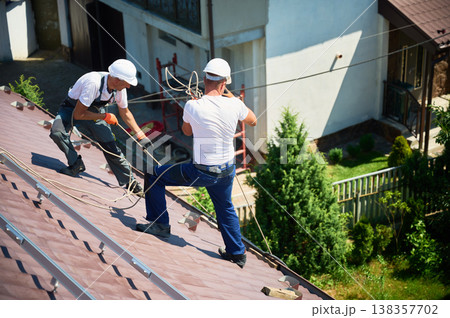 Men workers installing solar panel system on roof of house. Electricians in helmets lifting up photovoltaic solar module with help of ropes outdoors. Concept of alternative and renewable energy. Men workers installing solar panel system on roof of house. Electricians in helmets lifting up photovoltaic solar module with help of ropes outdoors. Concept of alternative and renewable energy. 138357702