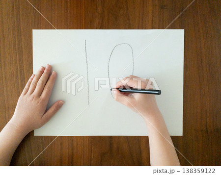 白い紙に黒ペンで数字の10を書く子どもの手　木のテーブルの上で学習する様子 138359122