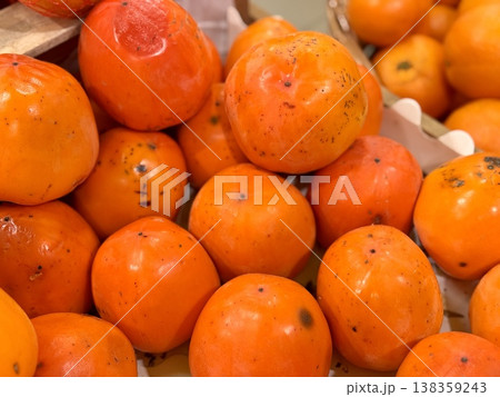 A pile of ripe juicy orange persimmons in a box, offered for sale at a supermarket vegetable stand, demonstrates organic, vegetarian and healthy food. Close-up. A pile of ripe juicy orange persimmons in a box, offered for sale at a supermarket vegetable stand, demonstrates organic, vegetarian and healthy food. Close-up. 138359243