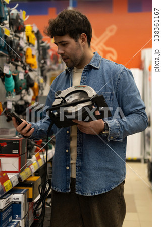 Man holding circular saw and smartphone, researching tools in hardware store Man holding circular saw and smartphone, researching tools in hardware store 138361167