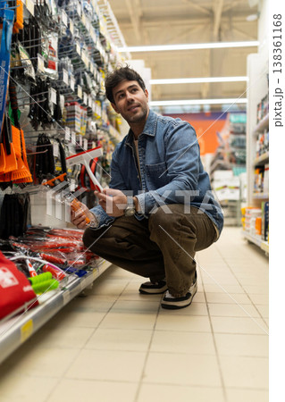 Young man shopping for household supplies, selecting a window squeegee from a shelf Young man shopping for household supplies, selecting a window squeegee from a shelf 138361168